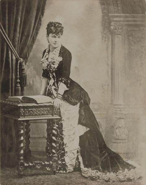 La marquise, en robe fin XIXe siècle, debout regardant vers à la droite, et s'appuyant sur une table où se trouve un livre.