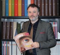 Portrait de Fabien Simode devant une bibliothèque, tenant un exemplaire du magazine L'OEil